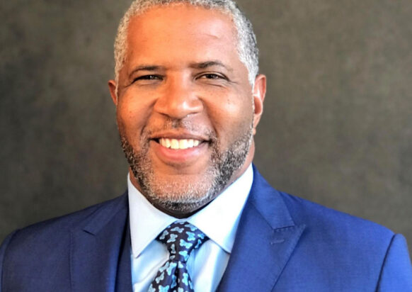 Robert F. Smith smiles while wearing a white shirt, patterned tie and blue suit and vest in front of a gray background